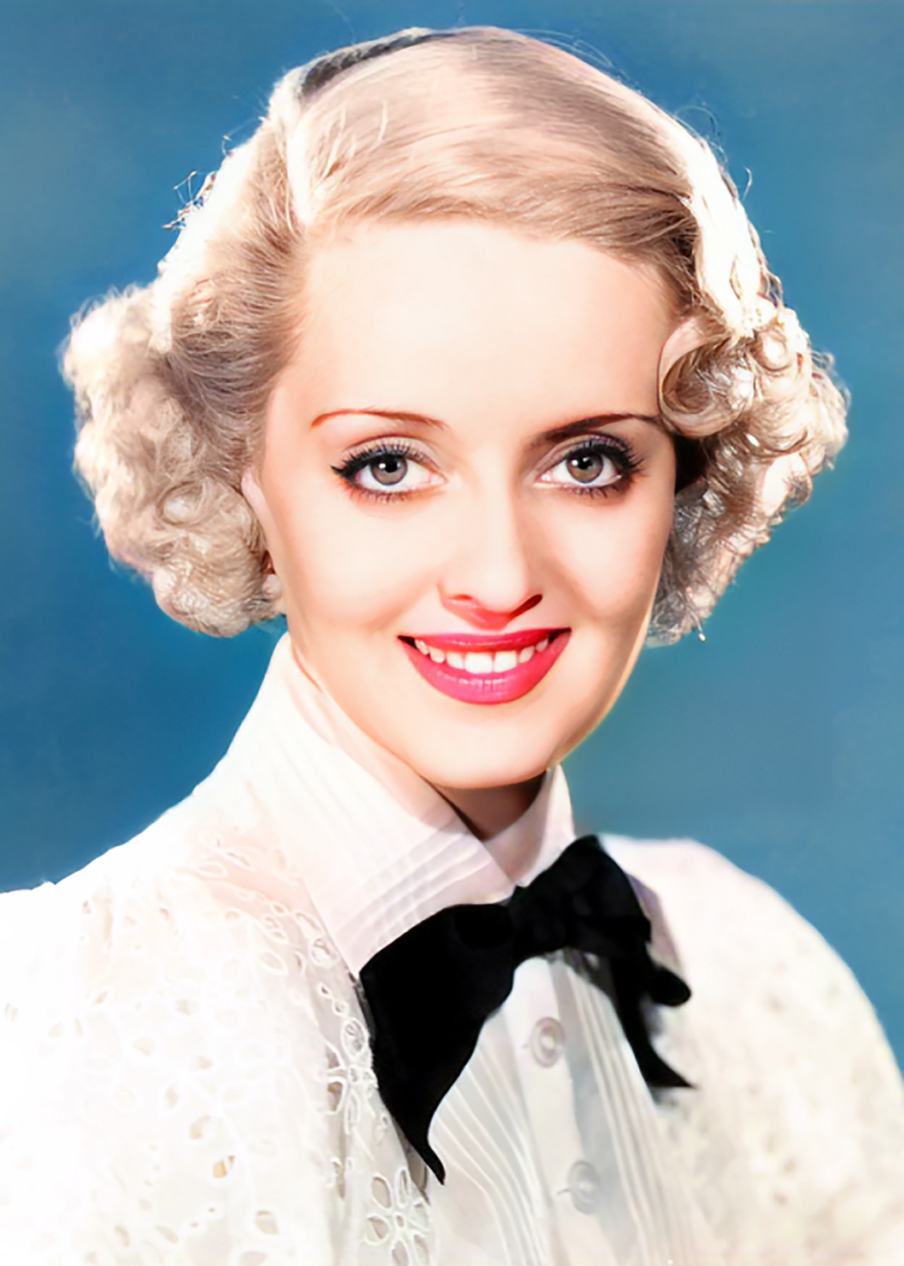 Colour-treated studio portrait of Bette Davis in 1935, wearing a white blouse with a black bow.