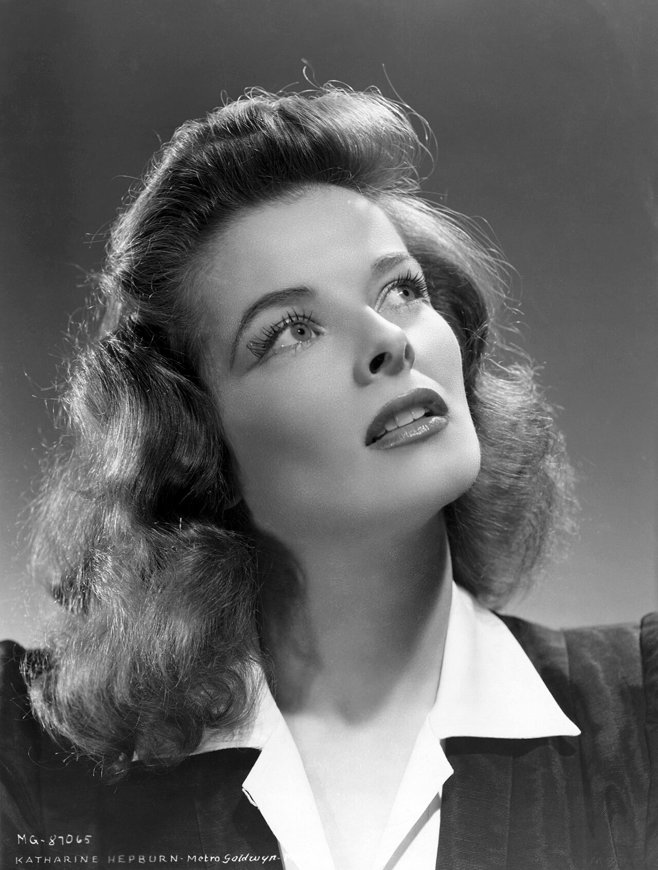 Black-and-white studio portrait of Katharine Hepburn looking upward, with swept-back hair and a white-collared jacket.