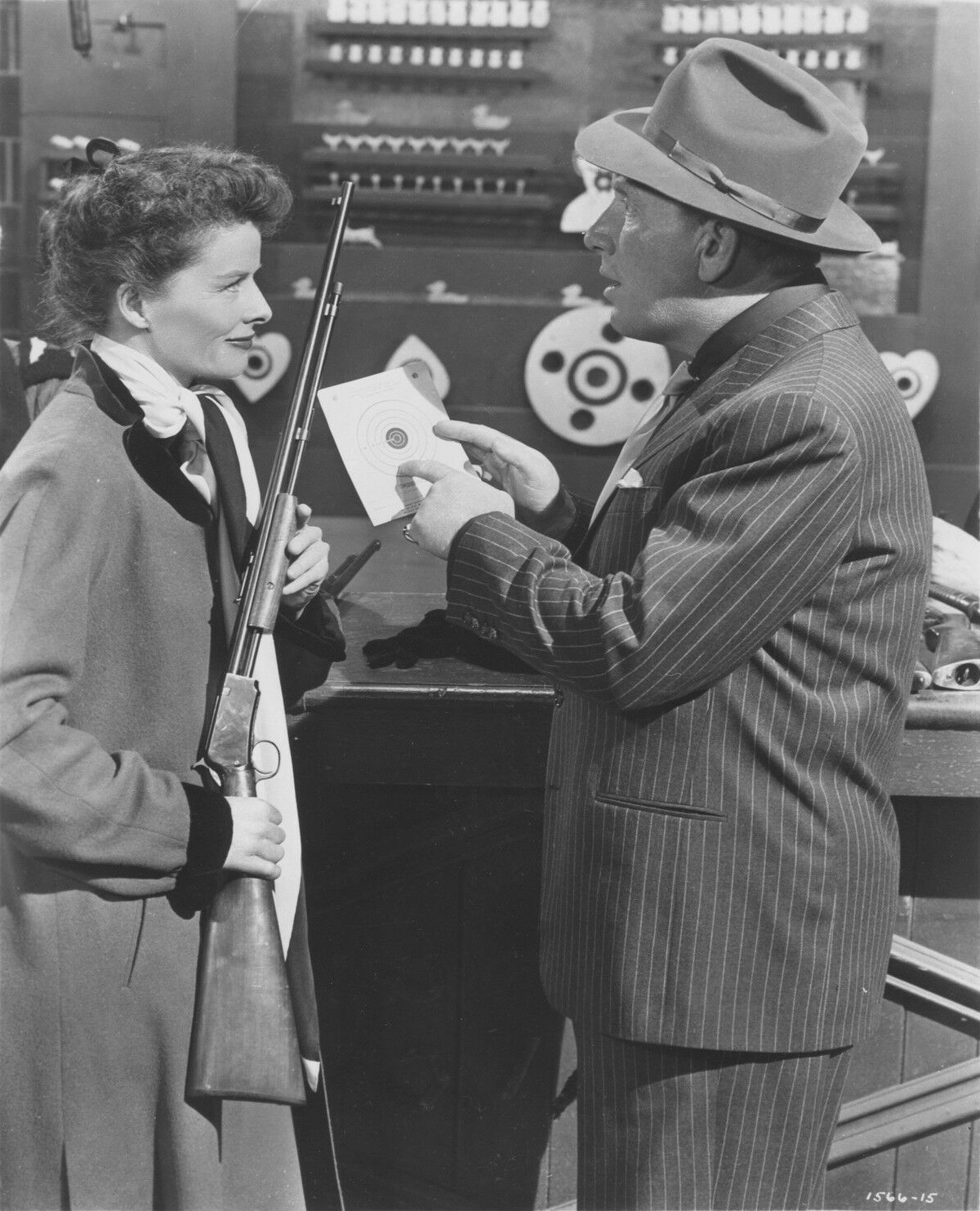 Katharine Hepburn holds a prop rifle while Spencer Tracy speaks to her beside a shooting target in Pat and Mike.