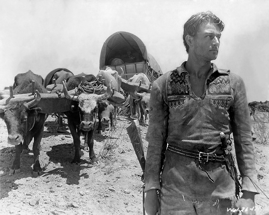 Young John Wayne stands outdoors in cowboy costume in a black-and-white publicity photograph for The Big Trail.